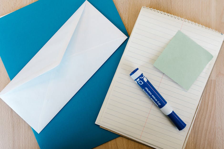 Flat lay of office supplies on a wooden desk, including a notepad, envelope, marker, and sticky note.